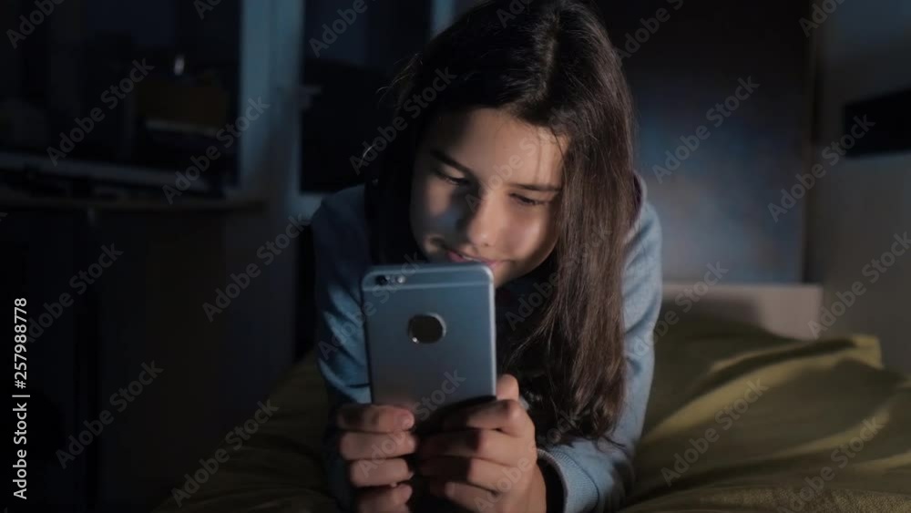 Teenage girl online shopping looks on her smartphone in bed during the night. little teen hood girl writes a message chatting in social media messenger night evening indoors smartphone on the bed Teenage girl online shopping looks on her smartphone in bed during the night. little teen hood girl writes a message chatting in social media messenger night evening indoors smartphone on the bed