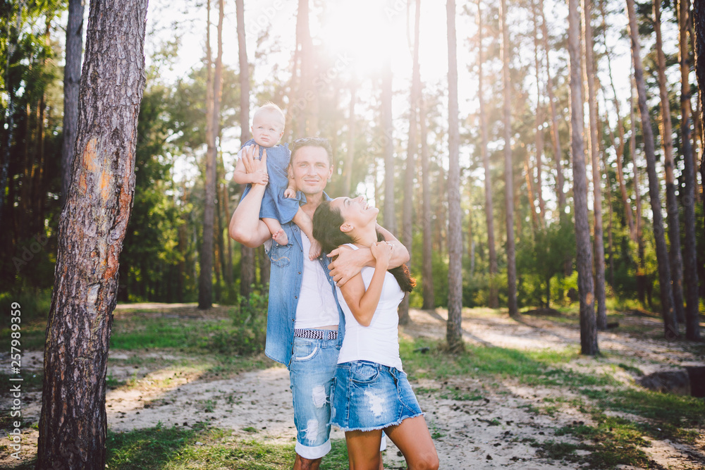 Fototapeta premium Family mom, dad and daughter sits at the daddy on the shoulders, and parents kiss on the nature in the forest in the summer at sunset
