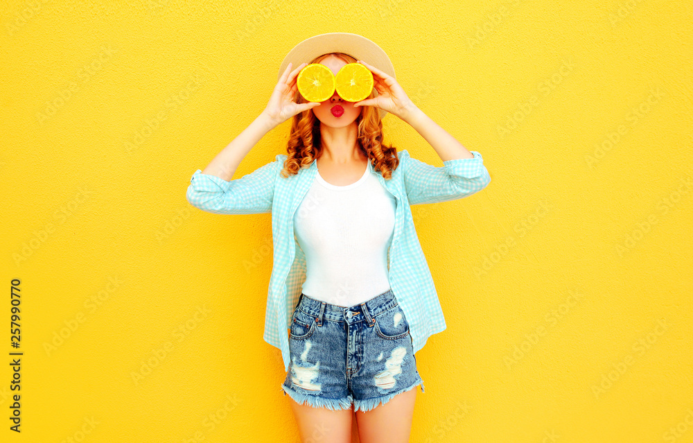 Summer portrait happy woman holding in her hands slices of orange hiding her eyes in straw hat, shorts on colorful yellow background Summer portrait happy woman holding in her hands slices of orange hiding her eyes in straw hat, shorts on colorful yellow background