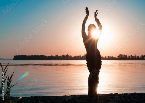 dark silhouette of dancing slim woman near big river coast at dawn