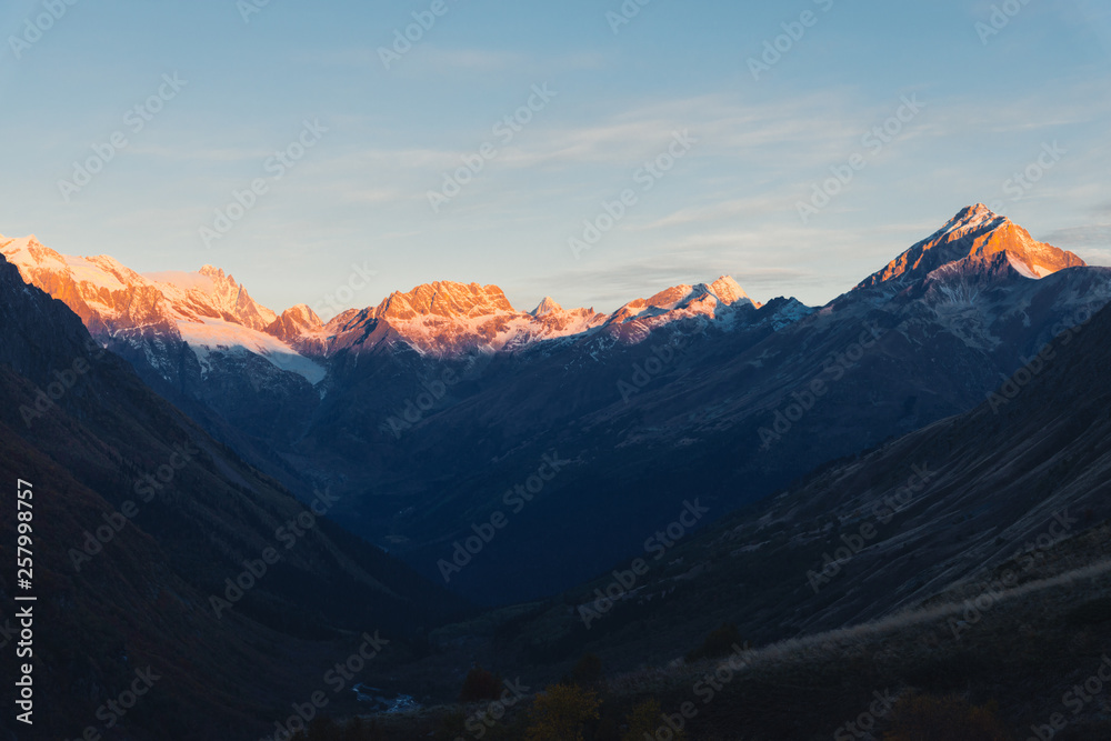 Sunset in the mountains. The snow-capped mountains are illuminated by the orange light of the setting sun, the valley below is in deep shadow. Copy space for text
