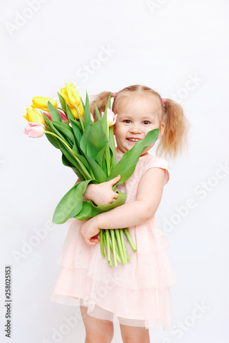 Little beautiful girl blonde with a bouquet of tulips on a light background. Baby girl smiling. Spring and women s day concept. Little girl holding a bouquet of colored tulips.   opy spase