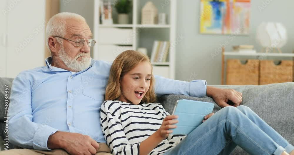 Portrait shot of the excited caucasian grandfather and granddaughter watching video on the tablet device while sitting together on the sofa in the cozy living room. Portrait shot of the excited caucasian grandfather and granddaughter watching video on the tablet device while sitting together on the sofa in the cozy living room.