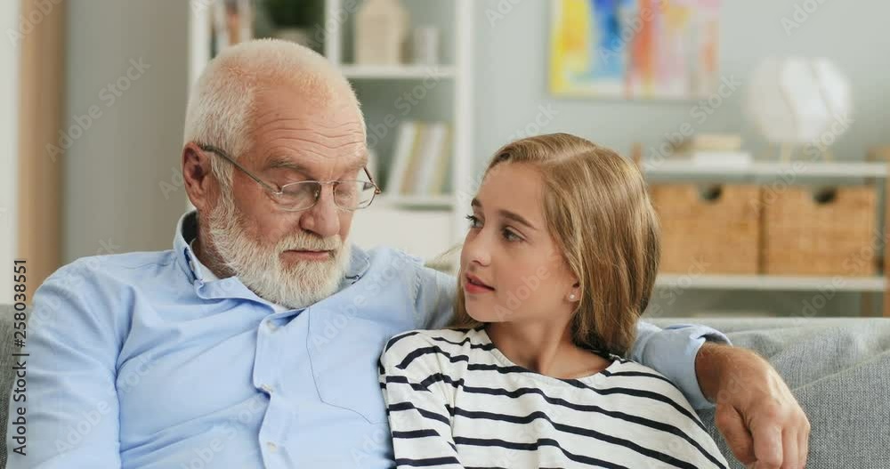 Portrait of the smiled Caucasian grandpa in glasses and blonde pretty grandaughter sitting close together on the sofa in the cozy room. Portrait of the smiled Caucasian grandpa in glasses and blonde pretty grandaughter sitting close together on the sofa in the cozy room.