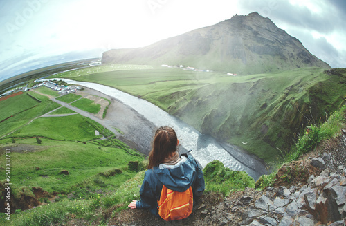 woman traveler in waterproof clothing sits on the cliff on background of Skogafoss waterfall in Iceland. View from above photo