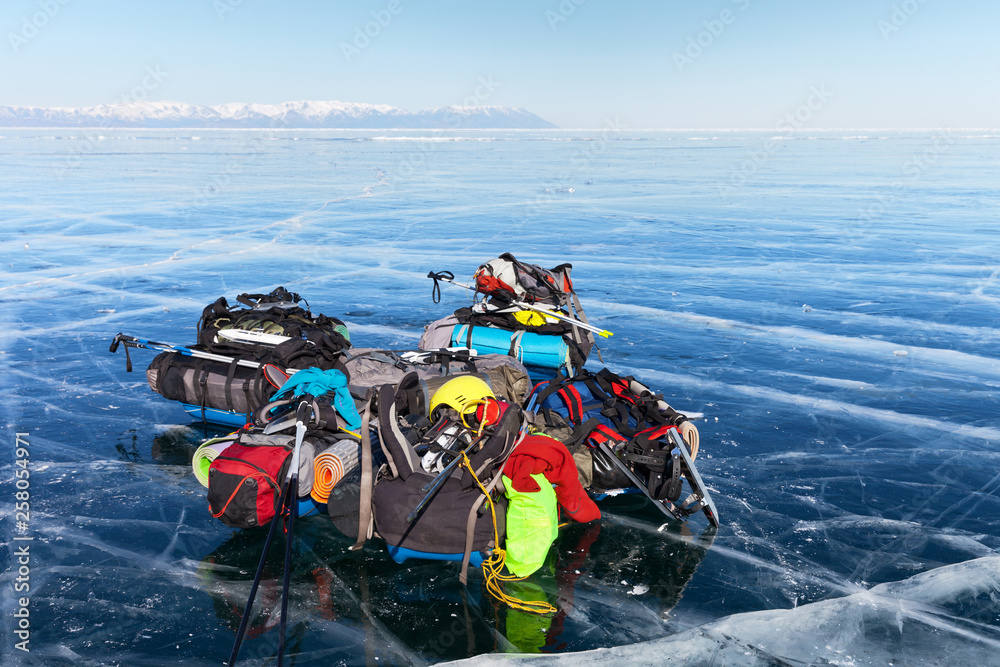 Obraz premium A pile of tourist equipment: tents, backpacks, mats, protective helmets, skates on beautiful blue ice against the boundless ice expanses of the frozen Lake Baikal. Winter tourism concept