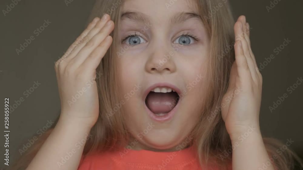 Cute little girl open her face with hands. Girl looks and is surprised and happy to receive a surprise. Portrait little young girl with blue eyes looking at camera. Closeup Cute little girl open her face with hands. Girl looks and is surprised and happy to receive a surprise. Portrait little young girl with blue eyes looking at camera. Closeup