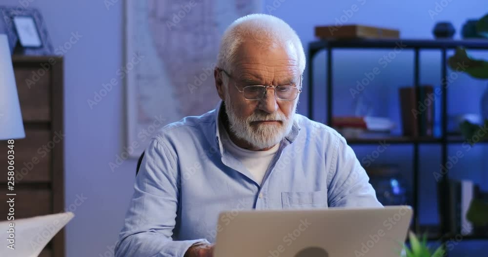 Portrait shot of the old man texting on the laptop computer and considering about his work while sitting at the desk late in the evening. Portrait shot of the old man texting on the laptop computer and considering about his work while sitting at the desk late in the evening.
