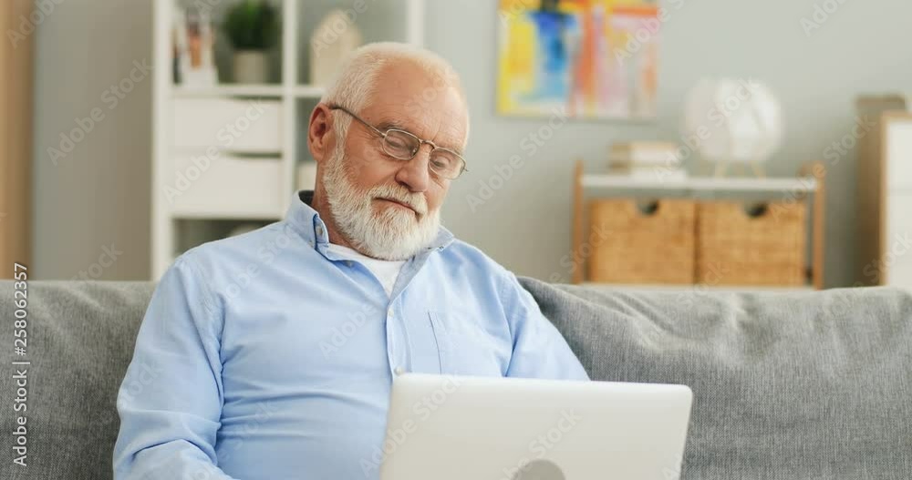 Senior man with grey hair and in glasses sitting on the couch, watching something on the laptop computer and smiling. Portrait. Senior man with grey hair and in glasses sitting on the couch, watching something on the laptop computer and smiling. Portrait.