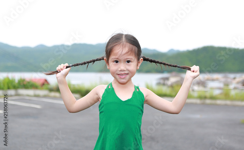 Happy little Asian child girl in the mountain field holding her pigtails hair with looking at camera.