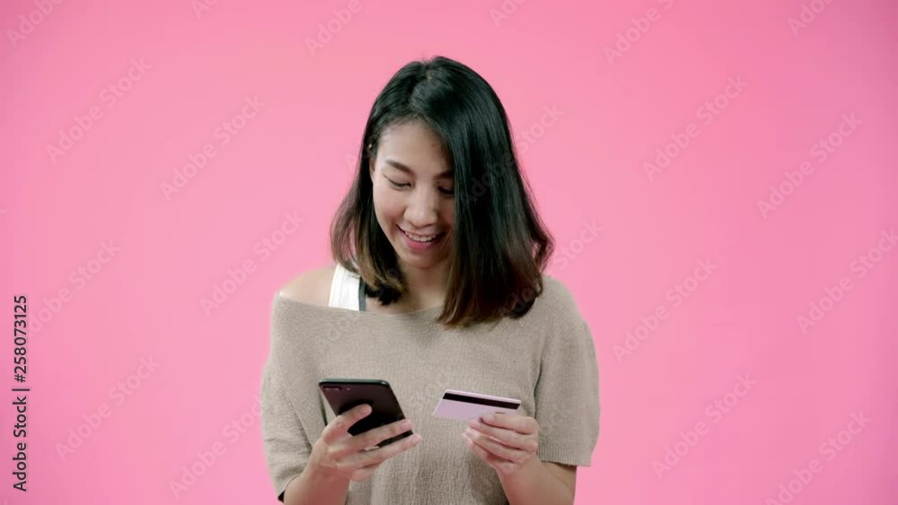 Young Asian woman using smartphone buying online shopping by credit card feeling happy smiling in casual clothing over pink background studio shot. Happy smiling adorable glad woman rejoices success. Young Asian woman using smartphone buying online shopping by credit card feeling happy smiling in casual clothing over pink background studio shot. Happy smiling adorable glad woman rejoices success.