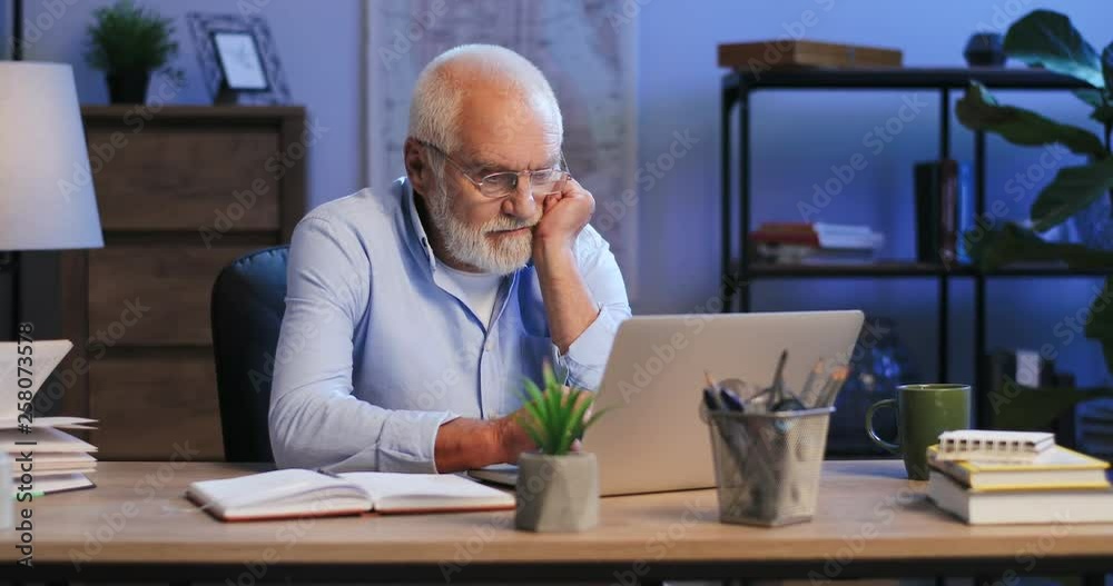 Senior Caucasian man in glasses sitting late at night at the laptop in his cabinet at home and thinking about work. Senior Caucasian man in glasses sitting late at night at the laptop in his cabinet at home and thinking about work.