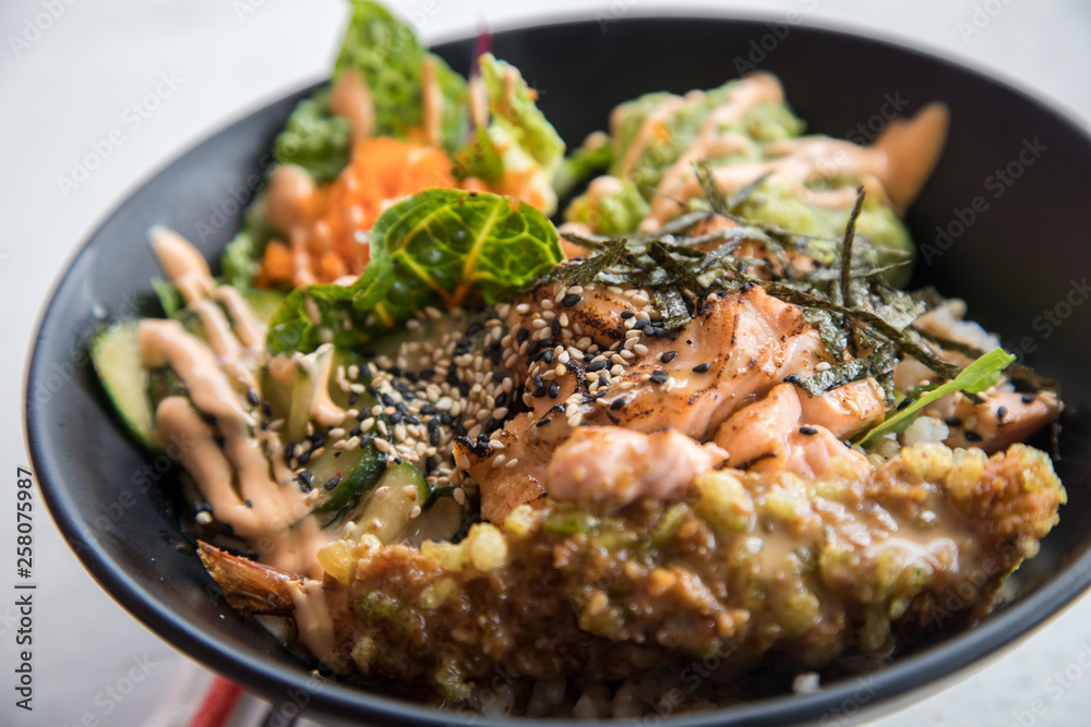 Poke Bowl mit flambierten Lachs, Tempura Shrimp, Guacamole, Masago Kaviar, Salat, Nori Algen und Sesam auf Reis in schwarz Porzellan Schale Poke Bowl mit flambierten Lachs, Tempura Shrimp, Guacamole, Masago Kaviar, Salat, Nori Algen und Sesam auf Reis in schwarz Porzellan Schale