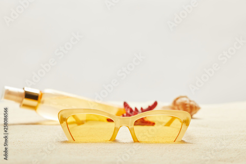 selective focus of yellow sunglasses near suntan oil bottle on golden sand isolated on grey