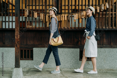 side view full length two asian women tourist visiting in japanese style wooden house with zen garden looking up enjoy nature spring view. young girls friends walk in walkway by wood hope cards wall