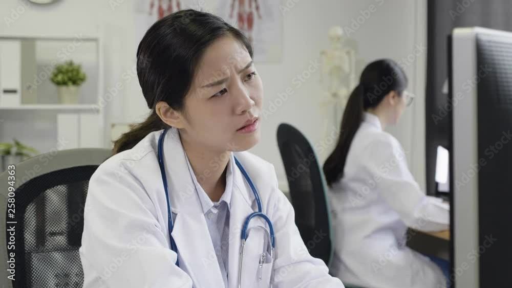 slow motion confused woman doctor sitting and using computer looking screen and document on desk frowning not understanding. colleague concentrated working in background in hospital office. slow motion confused woman doctor sitting and using computer looking screen and document on desk frowning not understanding. colleague concentrated working in background in hospital office.
