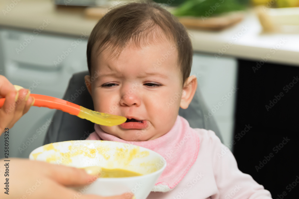 Mom feeds a baby with a spoon. The baby is crying while sitting at the baby’s feeding chair.