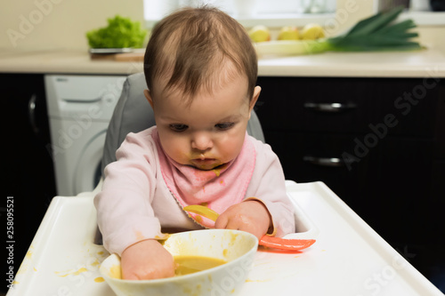 Feeding. Adorable baby child eating with a spoon in a children's chair. Baby's first solid food.