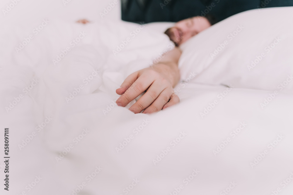 The hand of a man rests on white sheets. The man sleeps in a large bed of a hotel. It is located in a quiet and comfortable place. The hand of a man rests on white sheets. The man sleeps in a large bed of a hotel. It is located in a quiet and comfortable place.
