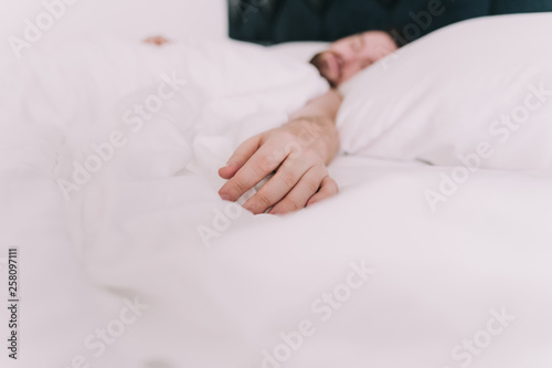 The hand of a man rests on white sheets. The man sleeps in a large bed of a hotel. It is located in a quiet and comfortable place.