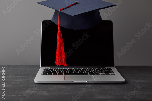 Laptop with blank screen and academic cap with red tassel on grey