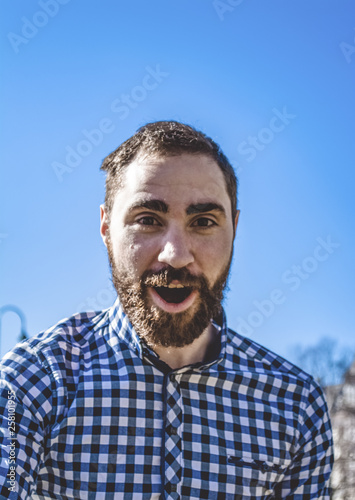 Portrait young bearded guy with emotions in shirt on the background of the blue clear sky in the park. Street fashion, hipster.