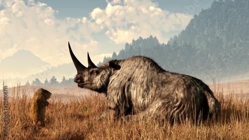 A marmot approaches an old and grey woolly rhinocerous. The great preshistoric rhino sits in long yellow grass with tree covered hills in the background. 3D Rendering