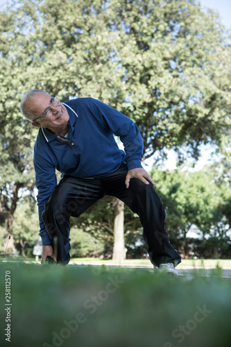 Uomo anziano con maglia blu sente un forte dolore alla caviglia in un parco