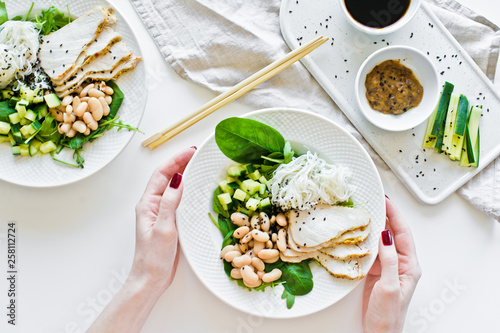 The girl holds a Buddha bowl with glass noodles, beans, chicken breast, spinach, arugula and cucumber. White background, top view