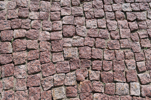 Paving stone laid out of square granite stones of brown color. Stones with jagged edges. The texture of the stones is mottled with dark dots. Day, the pavement lit by sunlight. Background, backdrop.