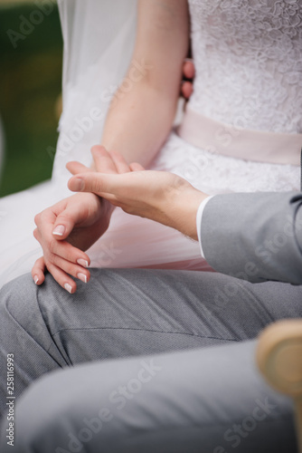 The bride and groom are sitting on a beautiful couch in a gazebo in the garden and holding hands. White tent. Green foliage. The bride in a pink fluffy dress  the groom in a gray suit.