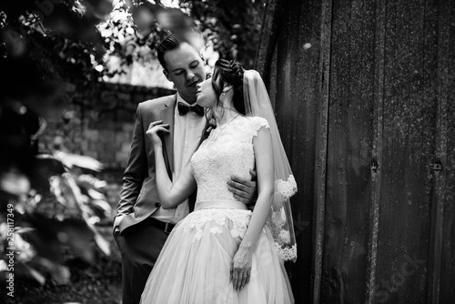 The bride and groom walk in the garden against the backdrop of a garland of light bulbs and metal doors. Black and white photo