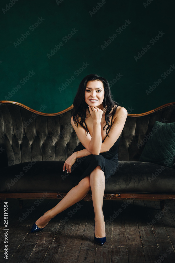 Beautiful girl in a black tight-fitting dress and heels. Black long wavy hair. Eastern appearance. Beautiful cut eyes. Brown sofa and green wall. A dark room. Sexy and stingy brunette. Portrait.