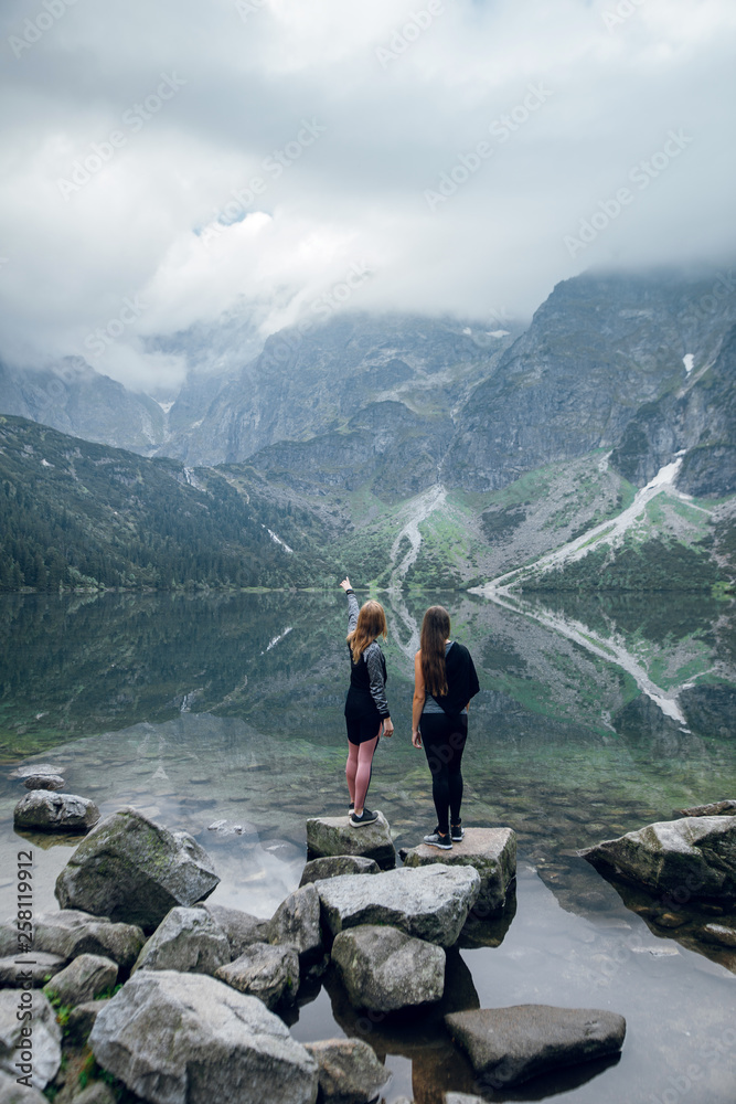 Two women, friends standing on the stony shore of lake Moskie Oko with scenic view of mountains with clouds and fog. Rysy mountains, Tatras. Poland, Slovakia. Rear view. Girl pointing on something