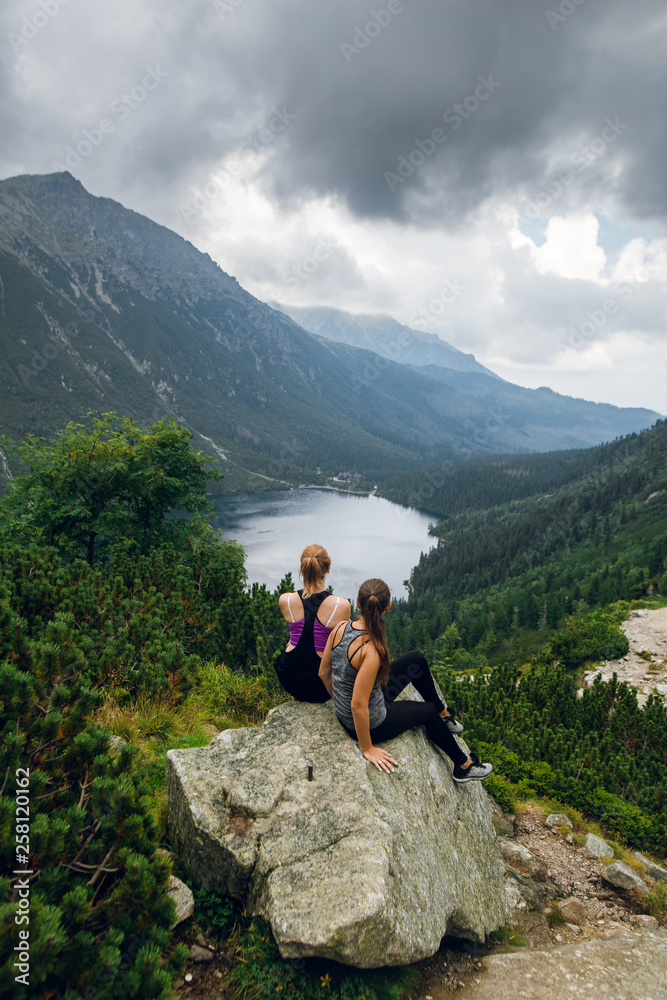 Two women friends sitting on the rocks, stones on the hill and admiring beautiful view of green hills and mountains on Morskie Oko lake, High Tatras, Zakopane, Poland. Top view