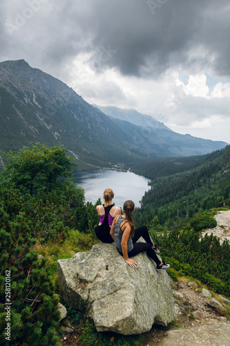 Two women friends sitting on the rocks, stones on the hill and admiring beautiful view of green hills and mountains on Morskie Oko lake, High Tatras, Zakopane, Poland. Top view