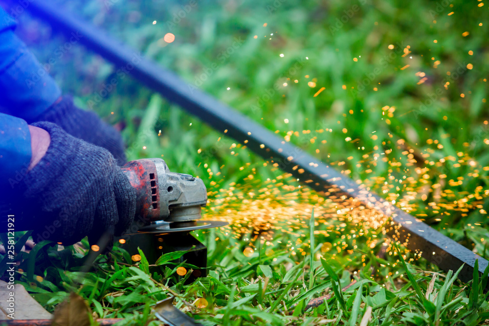 Naklejka premium hands of worker grinding or cutting metal with grinder at outdoor lawn or construction site