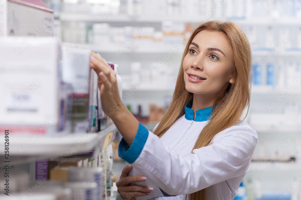 Beautiful confident female pharmacist smiling joyfully, organizing medical products on sale on the display. Professional chemist checking stock in an aisle, working at the drugstore. Medical industry 