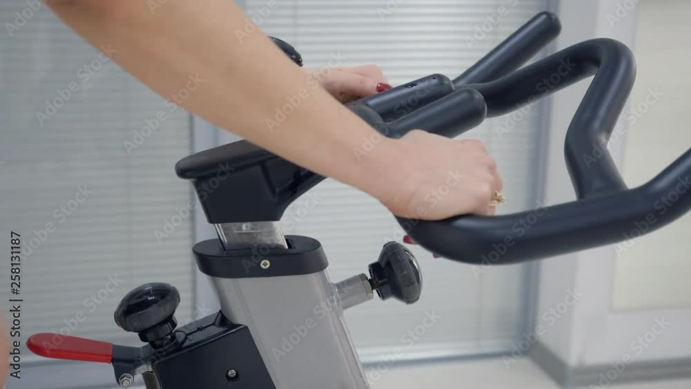 Hands of fitness woman holding handlebar indoor bike and adjusting force knob at bike in training gym. Cyclist woman using exercise bike for cardio training in fitness club. Sport lifestyle Hands of fitness woman holding handlebar indoor bike and adjusting force knob at bike in training gym. Cyclist woman using exercise bike for cardio training in fitness club. Sport lifestyle