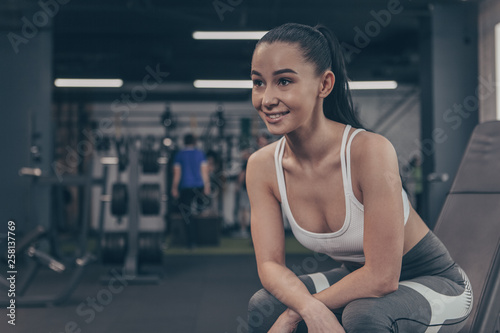 Attractive sportswoman smiling cheerfully, looking away, relaxing at the gym after intense workout. Happy female athlete in sportswear resting after exercising, copy space. Recreation, health concept