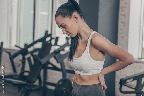 Beautiful athletic woman having backache after workout at the gym. Attractive fitness female having back or spine injury. Sportswoman rubbing her back, after tiring exercising. Injury, relief concept photo