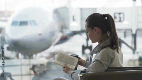 Beautiful traveler girl sits in airport lounge, drinks take away coffee and reads book on background of airplane in window