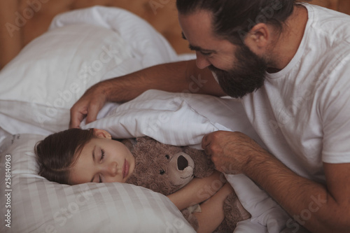 Loving father tucking blanket for his lovely little daughter. Adorable girl sleeping in bed, hugging her teddy bear, her father pulling up her blanket. Single parenting concept photo