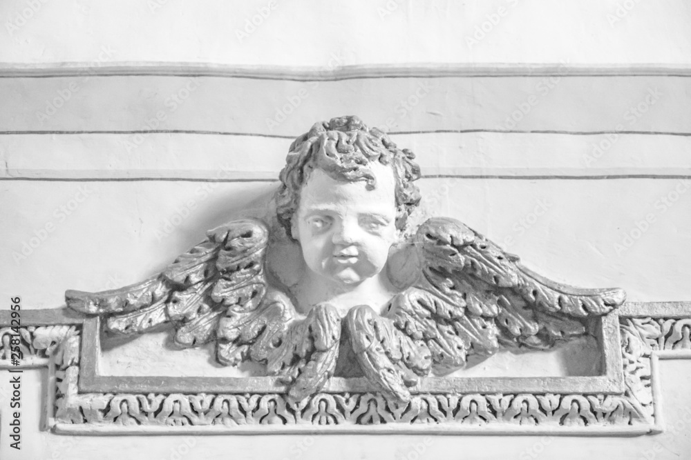 Fototapeta premium Closeup of an architectural detail. Bas-relief of a cherub with wings, enriched with stucco friezes, decorations, flowers and frames. Ancient sculpture attached to a plastered and painted wall.