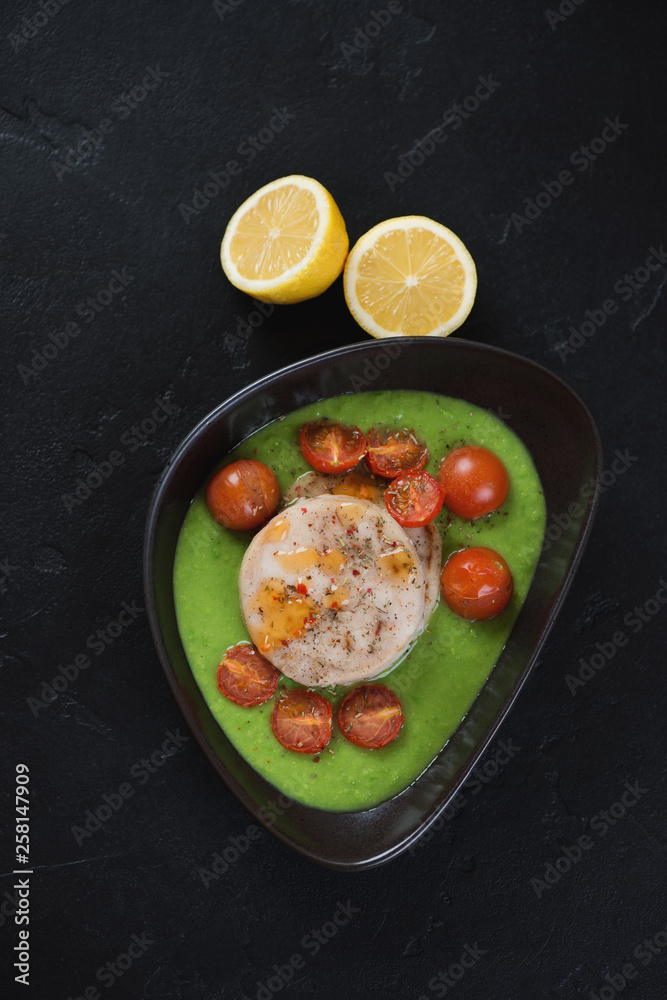 Bowl with baked hake medallions, green peas puree and cherry tomatoes. Flatlay on a black stone background, vertical shot