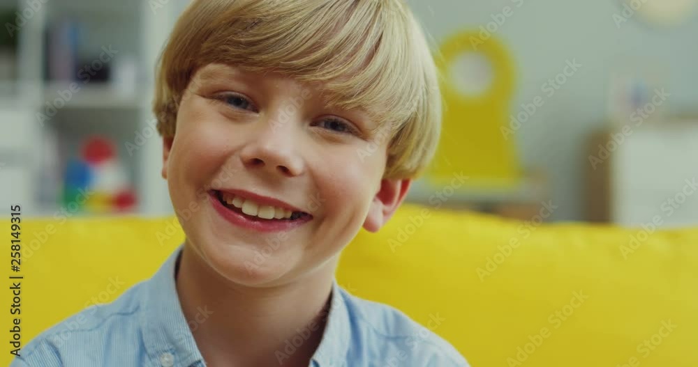Close up of the small blond teen boy turning his head to the camera and smiling cheerfully while sitting on the yellow couch in the living room. Portrait. Close up of the small blond teen boy turning his head to the camera and smiling cheerfully while sitting on the yellow couch in the living room. Portrait.