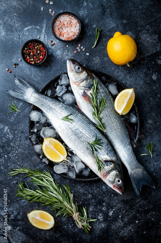 Fresh raw seabass fish on black stone background with spices, herbs, lemon and salt. Culinary seafood background with ingredients for cooking. Top view