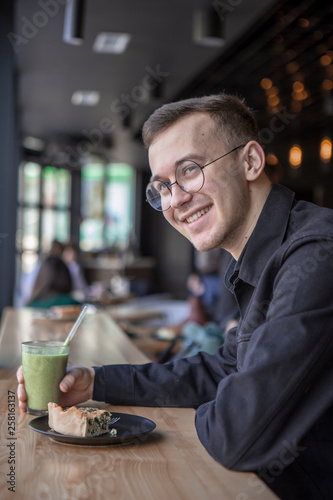 Young stylish man enjoys a cake and a drink while sitting in a cafe with large windows