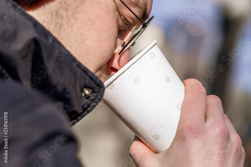 A stylish young boy, hipster, a student drinking coffee from a white paper cup strolling in a sunny day in the autumn or spring, glass with copy space for your text or logo