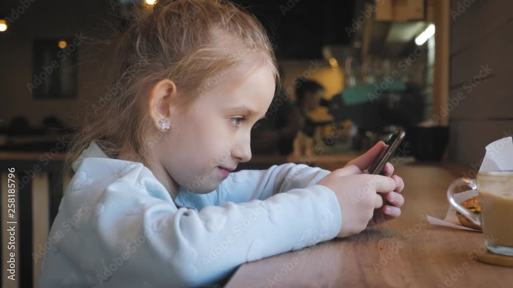 Cute little girl using on the smart phone at the cafe. Happy child having fun and relaxing after school. Natural indoor lifestyle. Cute little girl using on the smart phone at the cafe. Happy child having fun and relaxing after school. Natural indoor lifestyle.